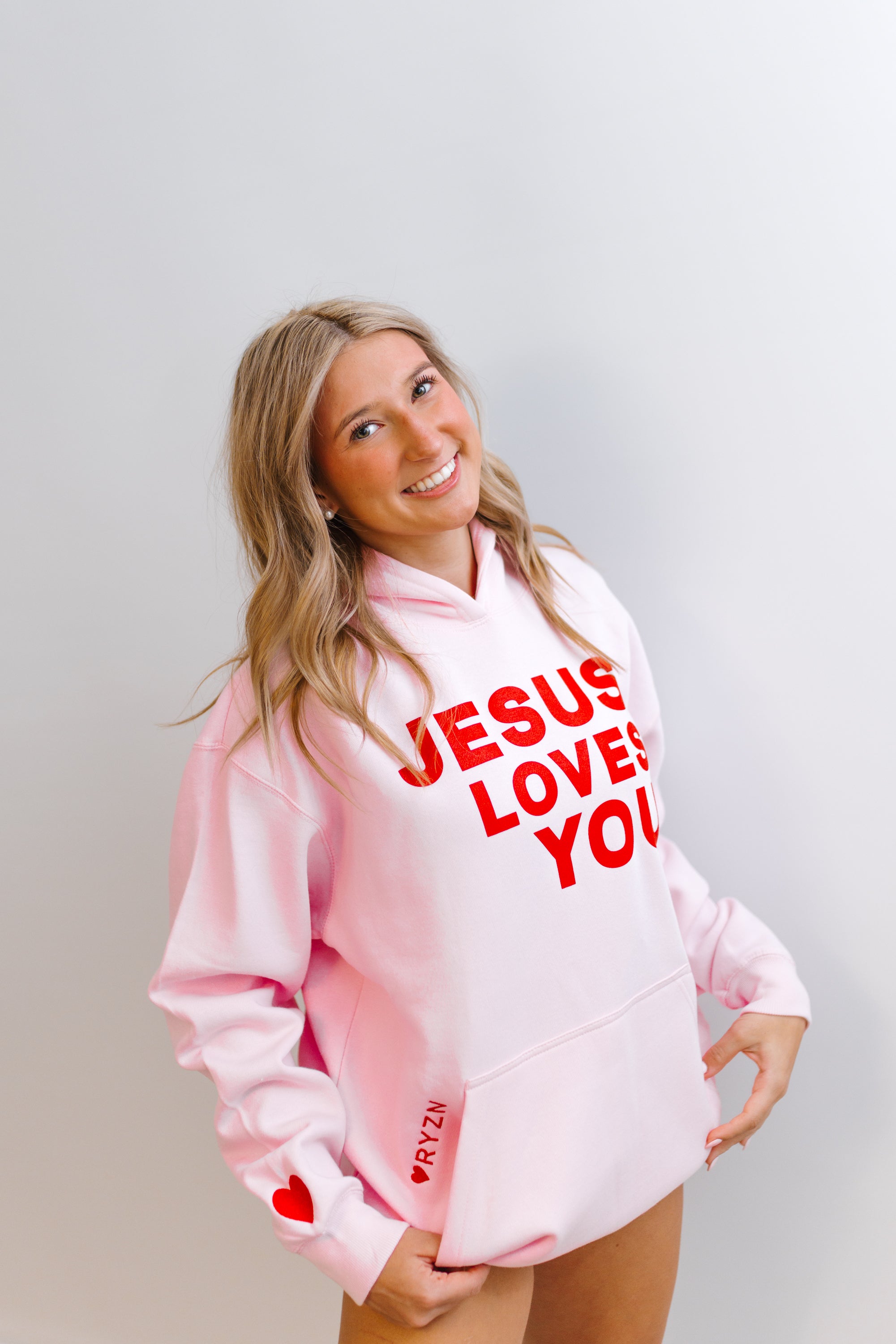Woman wearing a pink and white hoodie with 'Jesus Loves You' RYZN hoodie text on a plain background