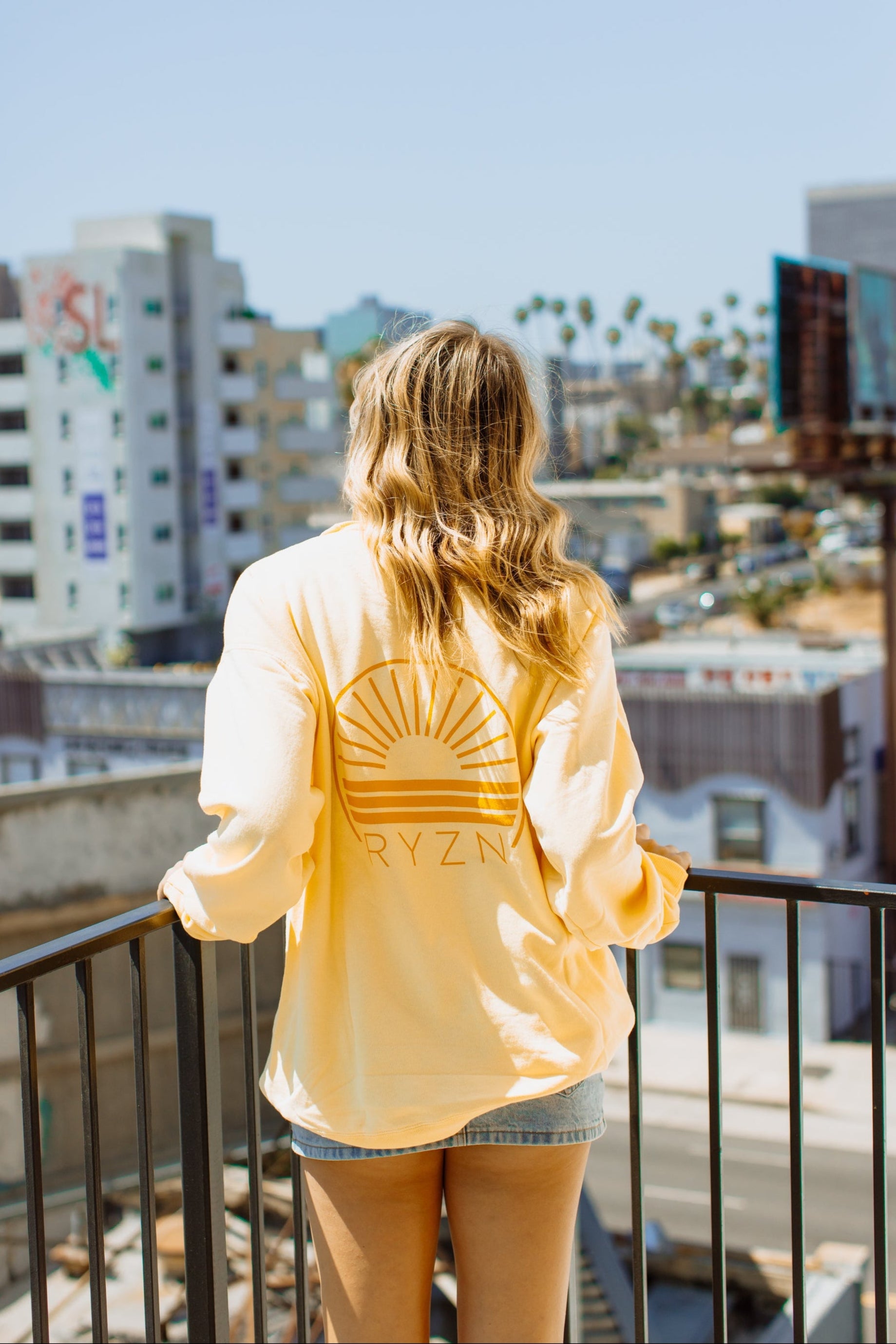 Person wearing a yellow shirt with a sun design and 'Ryzen' text, standing on a balcony overlooking a cityscape.